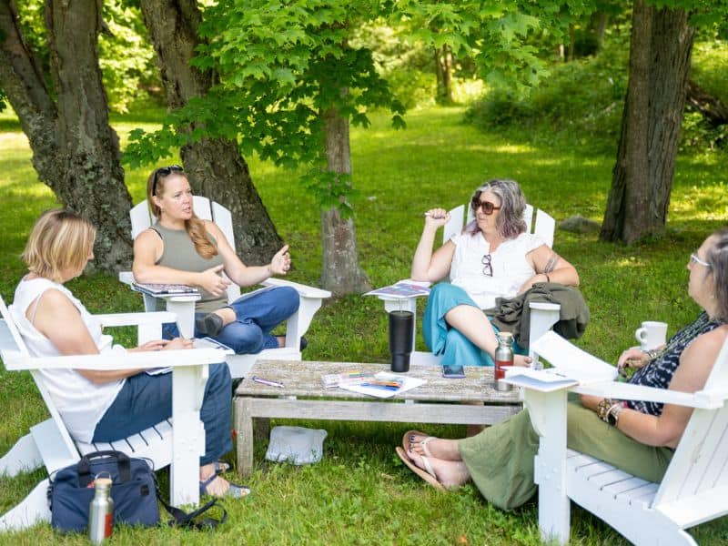 A group of writers sits in white Adirondack chairs around a small table on a grassy lawn surrounded by trees.
